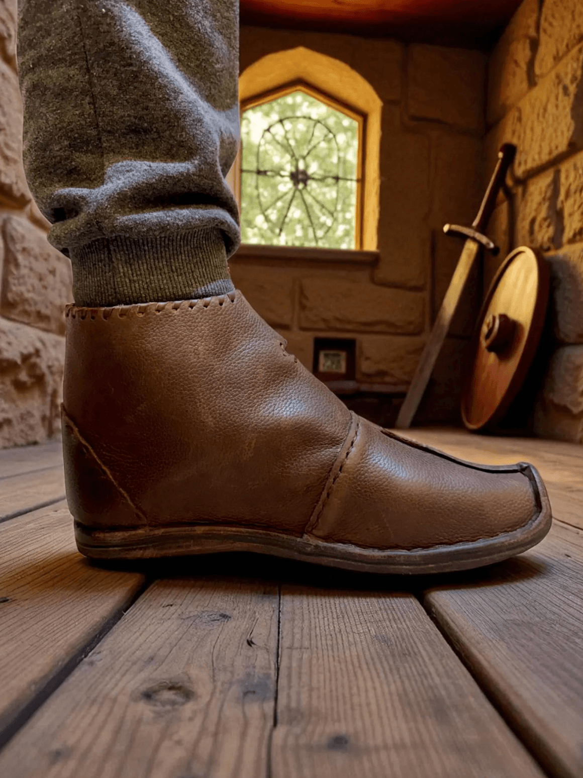 Handmade brown medieval leather boot with visible stitching, worn with gray pants in a stone room with sword and shield.